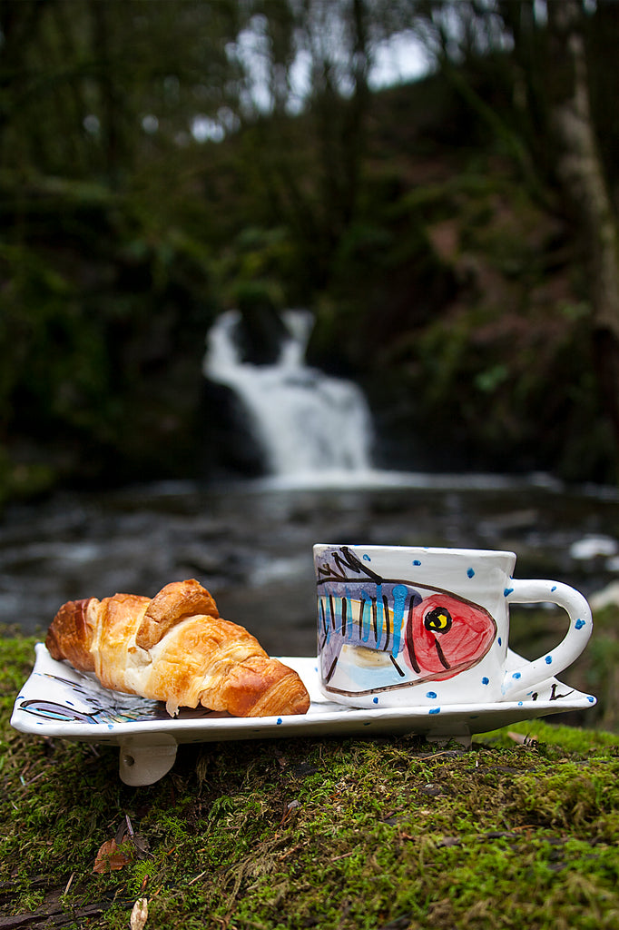 A handmade pottery mug rests atop a small rectangular plate. Each is crisp white with little blue dots hand-painted and smiling mackerel fish. They are atop a mossy stone with an Irish forest and waterfall behind. Handmade in Ireland.