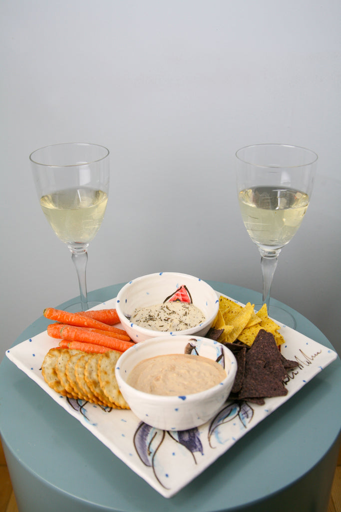 Showing how to use the ramekin bowl to serve dips. Two such ramekin bowls rest atop a medium or large square plate with crackers, carrot sticks, and tortilla chips. Two glasses of wine stand at the ready. Handmade in Ireland.