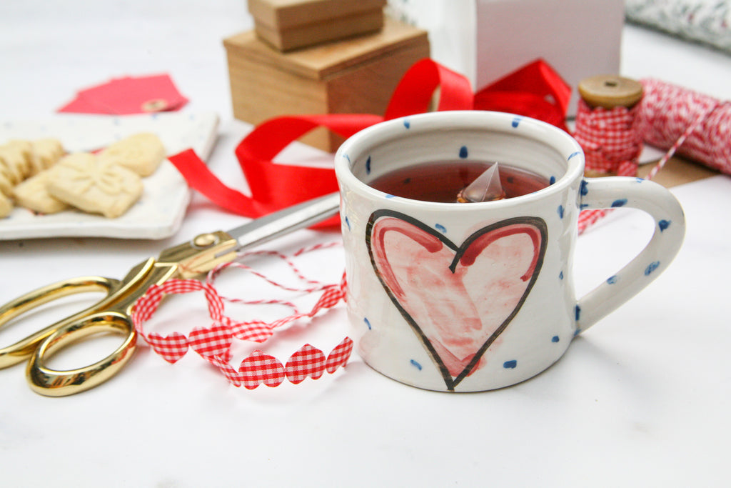 A handmade Irish pottery mug filled with tea sits beside sewing scissors and ribbon. A coordinating square small plate in the background holds cookies. Each is white with little blue dots and the focal point is a centred red heart with a black outline. Handmade in Ireland.