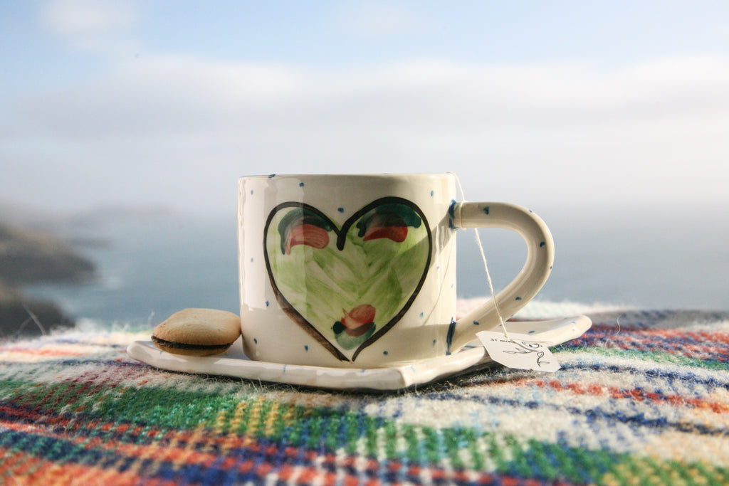 A handmade pottery mug rests atop a small square plate. Each is crisp white with little blue dots hand-painted and the focal point is a centred green heart with a black outline. They are atop a tartan blanket with the Irish countryside and coastline behind. Handmade in Ireland.