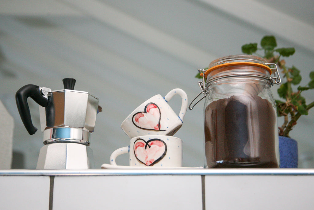 Two stacked hand painted red heart on a white ceramic mug with little blue dots. They sit between a clear glass jar of coffee grounds and a traditional stovetop coffee maker.
