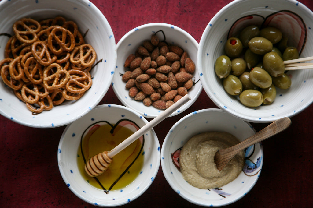 Showing how to use the ramekin bowl and small bowl sizes. Shown here with pretzels, almonds, olives, honey, and hummus in five different bowls. The white Irish ceramic pottery is peppered with blue dots and features the red heart design that is visible through the honey. Handmade in Ireland.