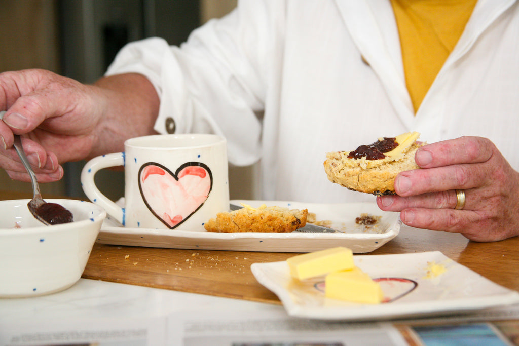 Person enjoying a dessert with a heart-themed mug and plates of food on a wooden table.