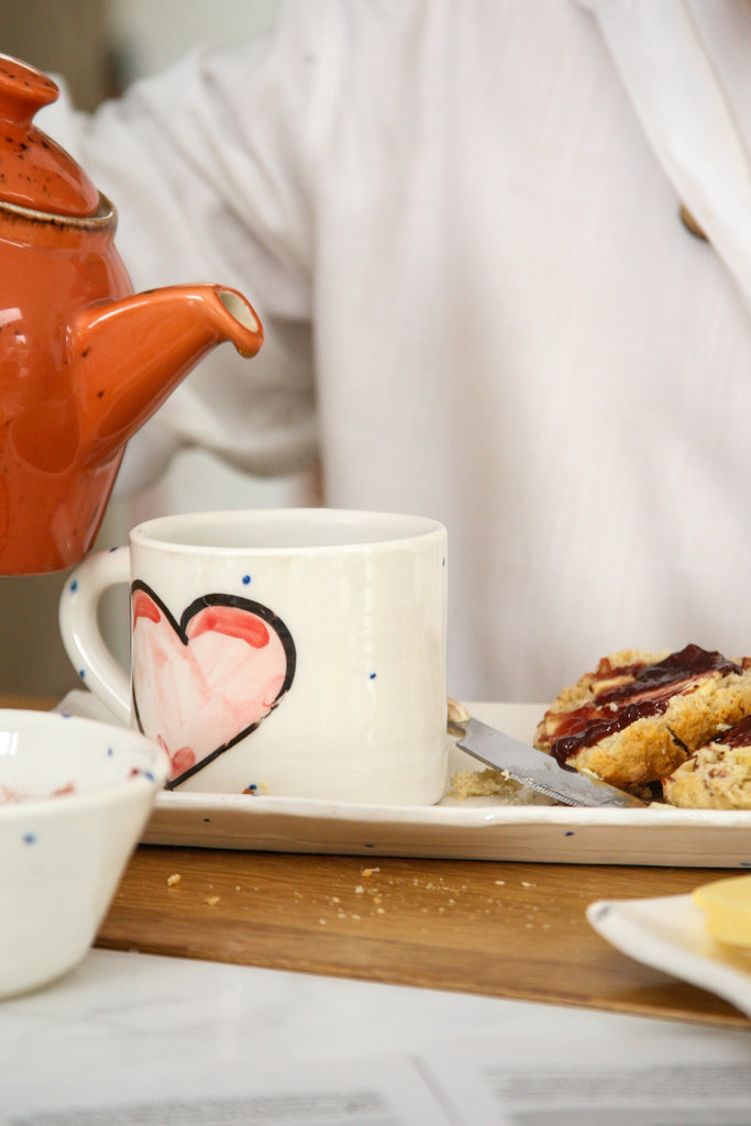 Mug of Tea with heart design, accompanying small bowl and plate , one teapot, with cookies on a wooden table.