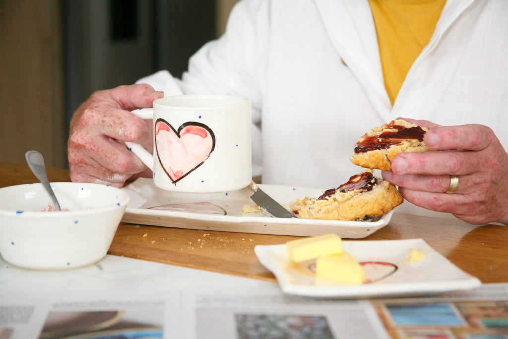 Person enjoying a meal with a mug featuring a heart design, scone, and other food items on a table.