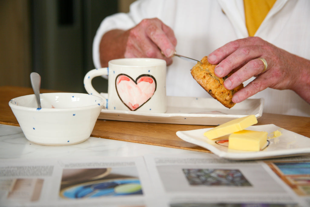 Person cutting a scone, buttering with butter, using knife and mug with serving plate and bowl.  All have  a Grá Heart design 