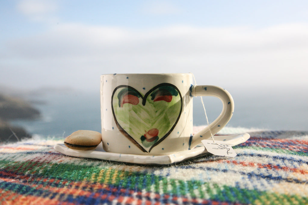 A handmade pottery mug rests atop a small square plate that acts as a saucer for the pottery mug. Each is crisp white with little blue dots hand-painted and the focal point is a centred green heart with a black outline.  Handmade in Ireland.