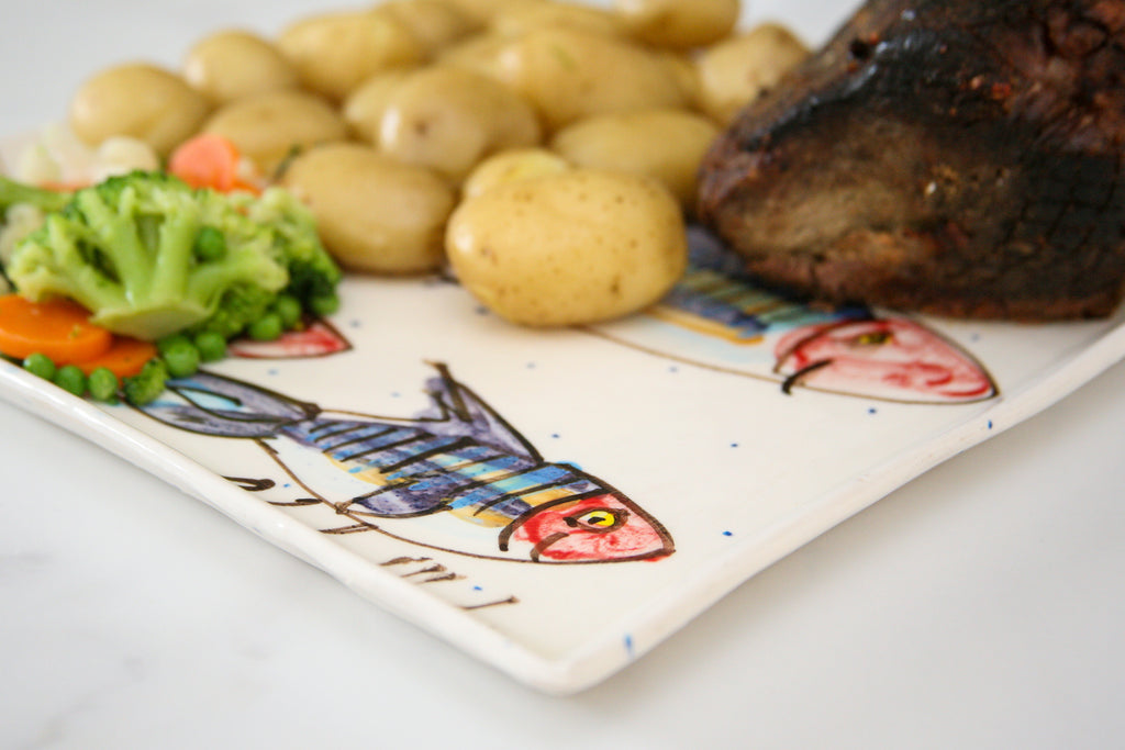 Large colourful fish-patterned plate with potatoes, broccoli, and carrots on a white surface