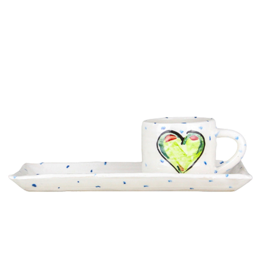 A white handmade pottery mug with blue dots and a big green heart resting atop a coordinating rectangular plate.