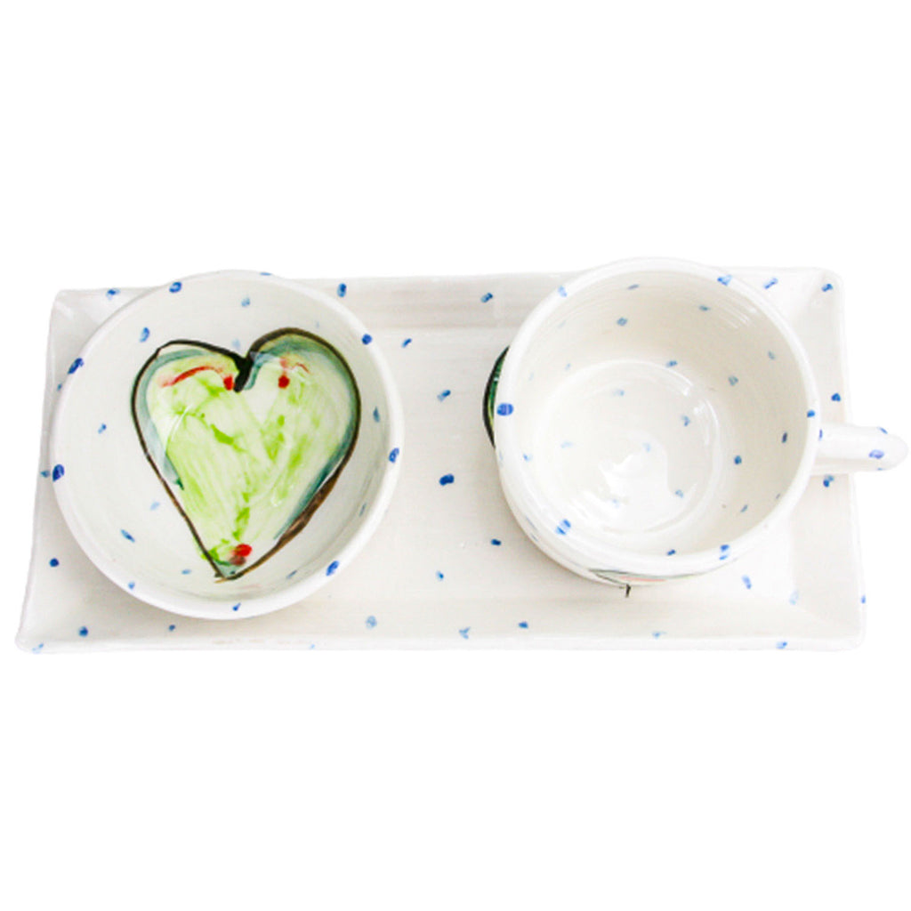 Aerial view of a white handmade pottery bowl with blue dots and a big green heart. It is resting alongside a coordinating mug and atop a coordinating rectangular plate.