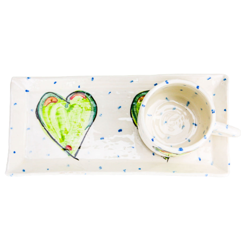 Aerial view of a white handmade pottery mug with blue dots resting atop a coordinating rectangular plate. Each is adorned with the handpainted green heart motif.
