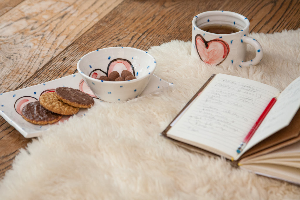 An open journal alongside a rectangular handmade pottery plate with cookies. A coordinating bowl rests atop the plate and the matching mug is beside it with tea. Each is handpainted with little blue dots and a red heart. The plate has two hearts. Handmade in Ireland.