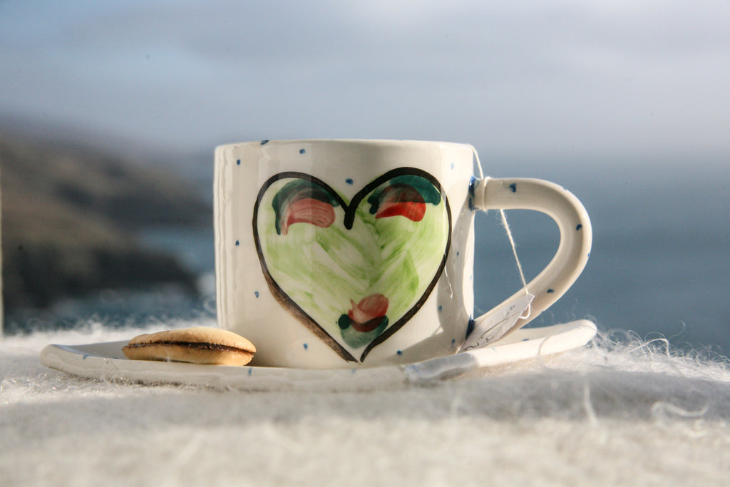 A handmade Irish pottery mug filled with tea. A coordinating square small plate is beneath the mug with a cookie. Each is white with little blue dots and the focal point is a centred green heart with a black outline. Handmade in Ireland.