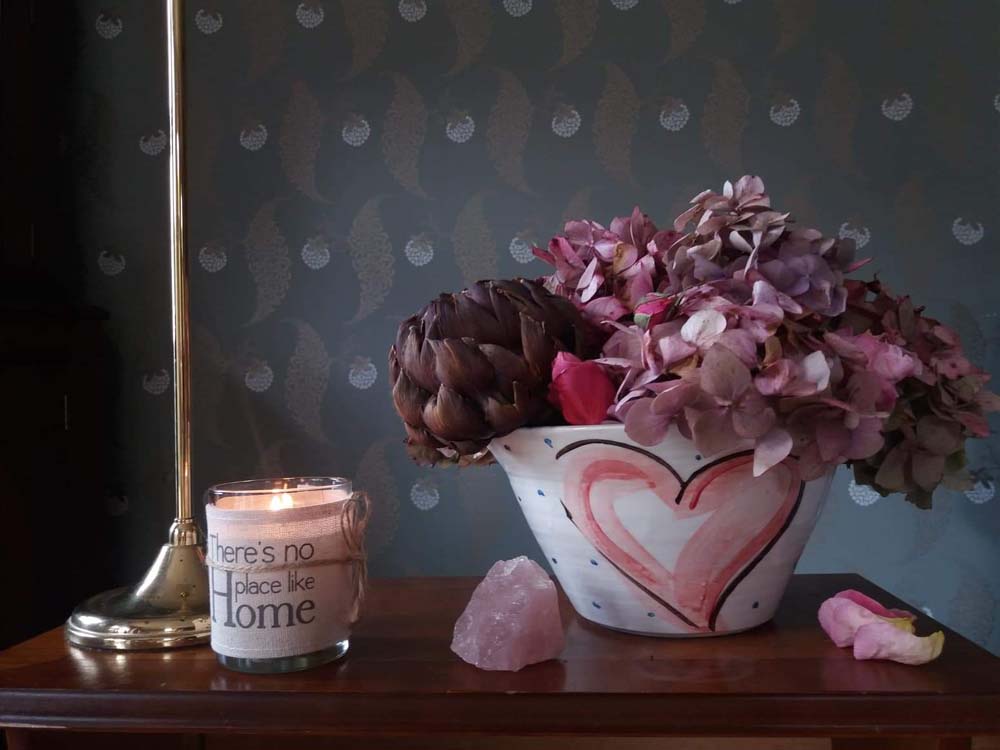 Large white ceramic bowl hand-painted with red hearts adorned with a black outline on this Irish pottery. Little blue dots pepper the high gloss finish. The bowl is filled with a bouquet of flowers, a mix of pink and lavender hydrangea and something like a dahlia. A candle is by its side. Handmade in Ireland.