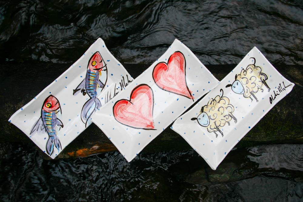 Three small rectangular platters side-by-side. The far left platter has two hand-painted mackerel fish on it. The middle platter has two red hearts outlined in black. The final plate on the right has two witty sheep hand-painted on it. Dark river water flows behind the platters.