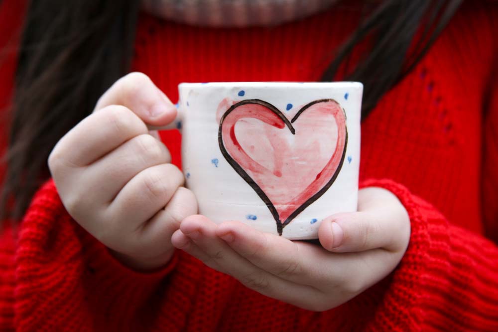 A hand holding the handle of a handmade Irish pottery mug. The mug is hand-painted with a red heart outlined in black. The holder is wearing a red sweater.