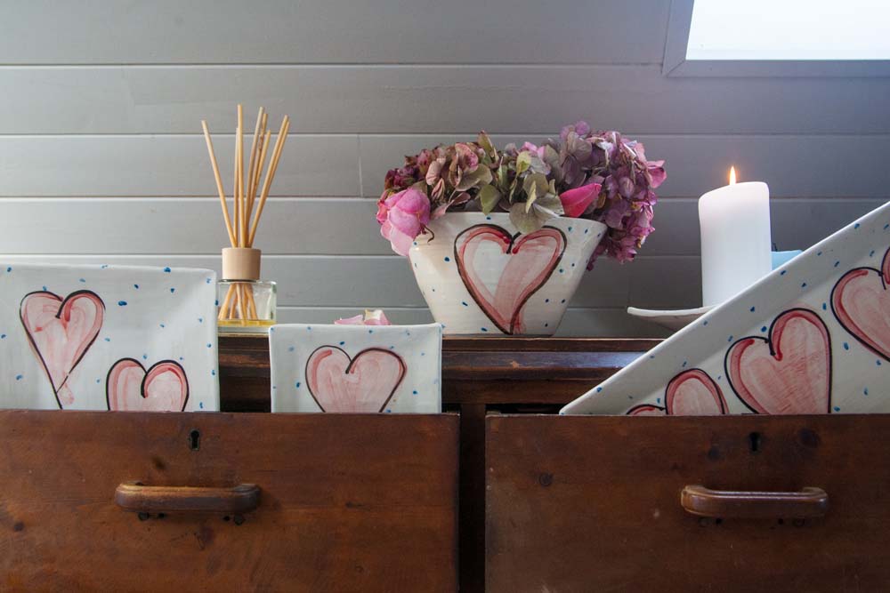 Red hand-painted hearts and little blue dots adorn each piece in the collection. Perfect for serving scones, snacks, breakfast, or tapas. Handmade in Ireland. Displayed here in a vintage wood dresser along with other pieces of the Grá Heart collection.