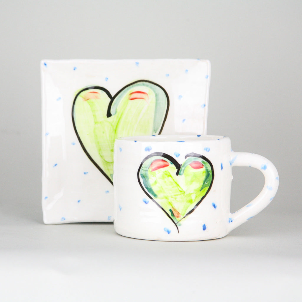 A handmade pottery mug sits in front of a square platter. Each is crisp white with little blue dots hand marked and the focal point is a centred green heart with a black outline. Handmade in Ireland.