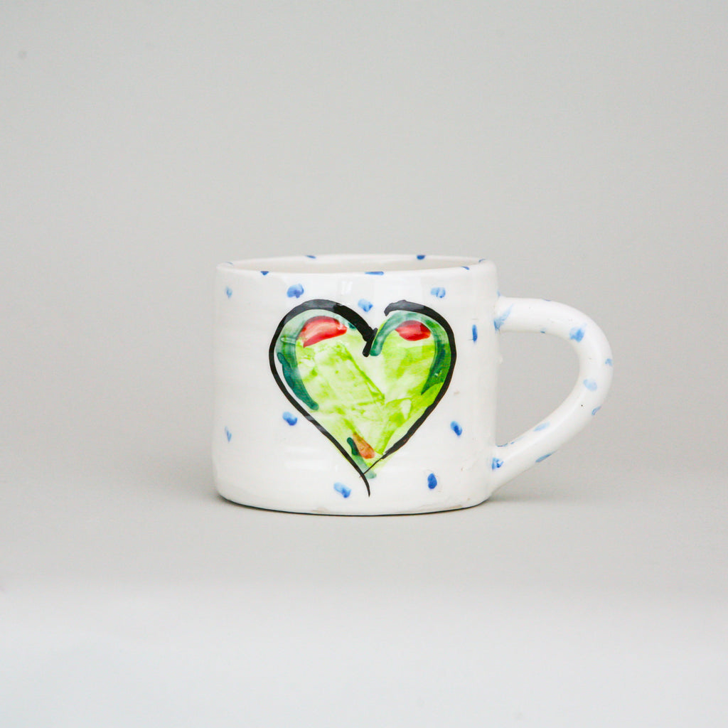 A handmade pottery mug. Crisp white Irish pottery with little blue dots hand marked and the focal point is a centred green heart with a black outline.