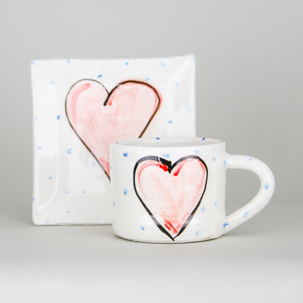 A handmade pottery mug in front of a small square plate. Each is crisp white with little blue dots hand marked and the focal point is a centred red heart with a black outline. Handmade in Ireland.