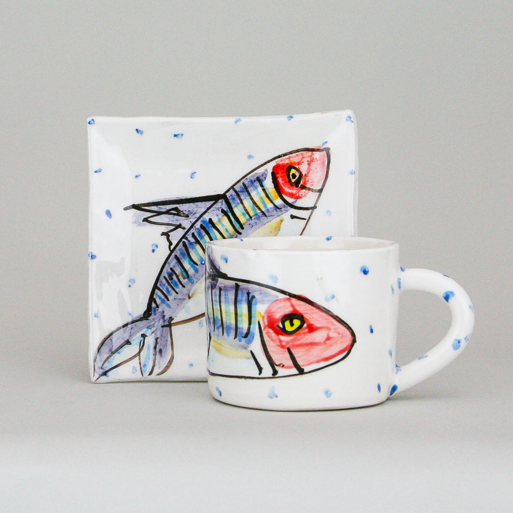 A handmade pottery mug in front of a square plate. Each is crisp white with little blue dots hand-painted along with a smiling mackerel fish. Handmade in Ireland.