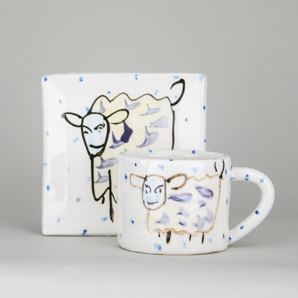A handmade pottery mug in front of a square plate. Each is crisp white with little blue dots hand-painted along with a witty sheep. Handmade in Ireland.