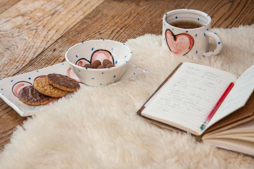 An open journal alongside a rectangular handmade pottery plate with cookies. A coordinating bowl rests atop the plate and the matching mug is beside it with tea. Each is handpainted with little blue dots and a red heart. The plate has two hearts. | Charlie Mahon Ceramics Pottery Cork Ireland 