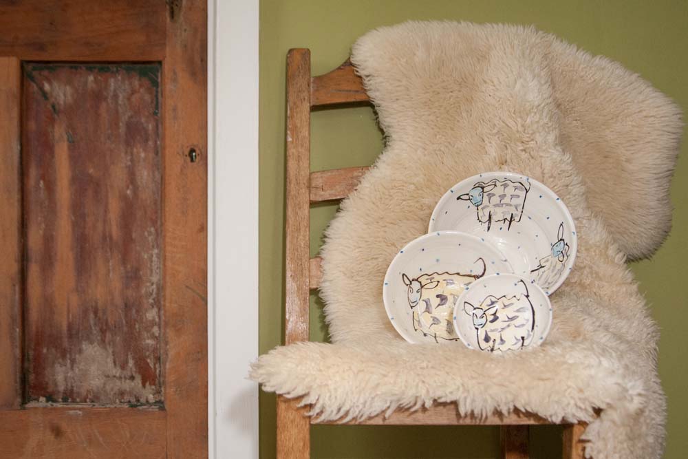Three progressively larger bowls on a sheepskin on a wood chair, each with hand-painted sheep surrounded by subtle blue dots . It is the small, medium, and large bowl sizes of the sheep collection. Handmade in Ireland.