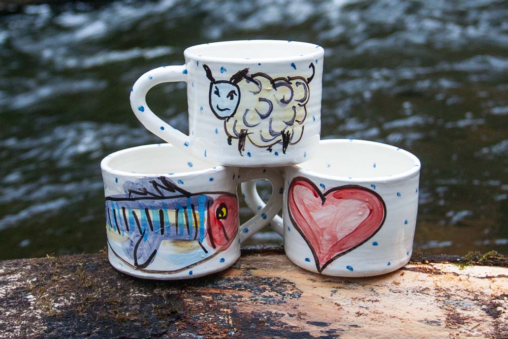 Trio of Irish pottery mugs on a large log in front of a river out of focus in the background. The mugs each have a different design, one with a handpainted sheep, one with a handpainted smiling mackerel, and the last with a red heart. Handmade in Ireland.