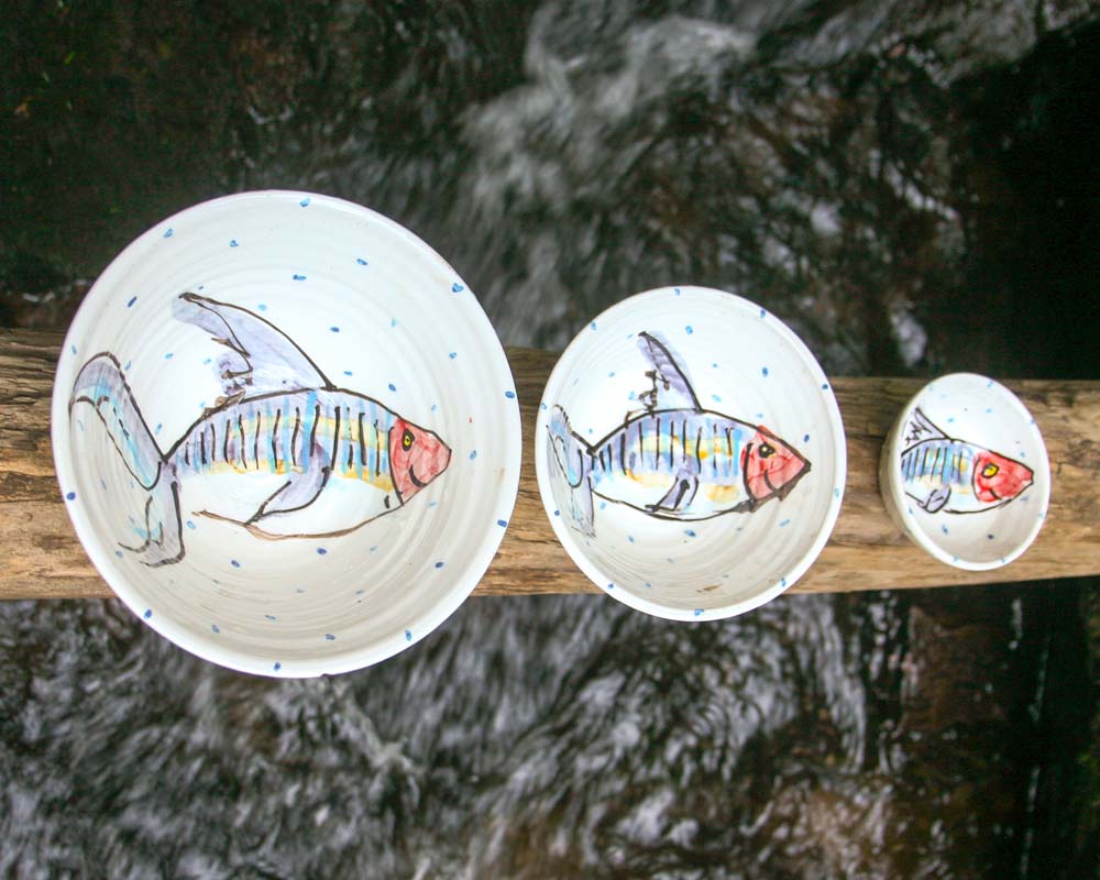 Three progressively larger bowls on a log, each with hand-painted blue and red mackerel fish in the centre. It is the small, medium, and large bowl sizes of the Mackerel fish collection. Handmade in Ireland.